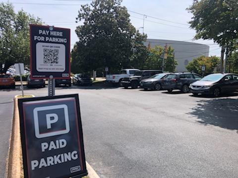A parking lot with several cars parked; signs in the foreground read "PAID PARKING" and "PAY HERE FOR PARKING" with a QR code for mobile payment. Trees and a building are visible in the background.