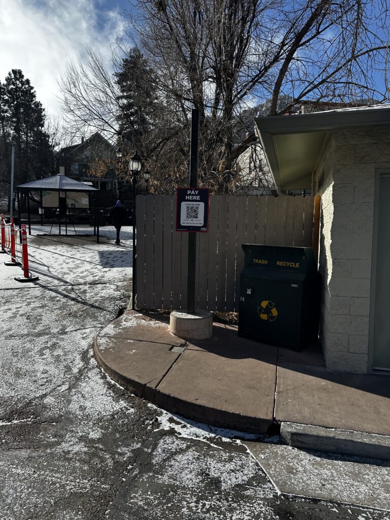 A snowy sidewalk curves past a recycling and trash bin near a small building. A sign with a QR code reads "Pay Here." In the background are trees, a pavilion, and orange traffic cones.