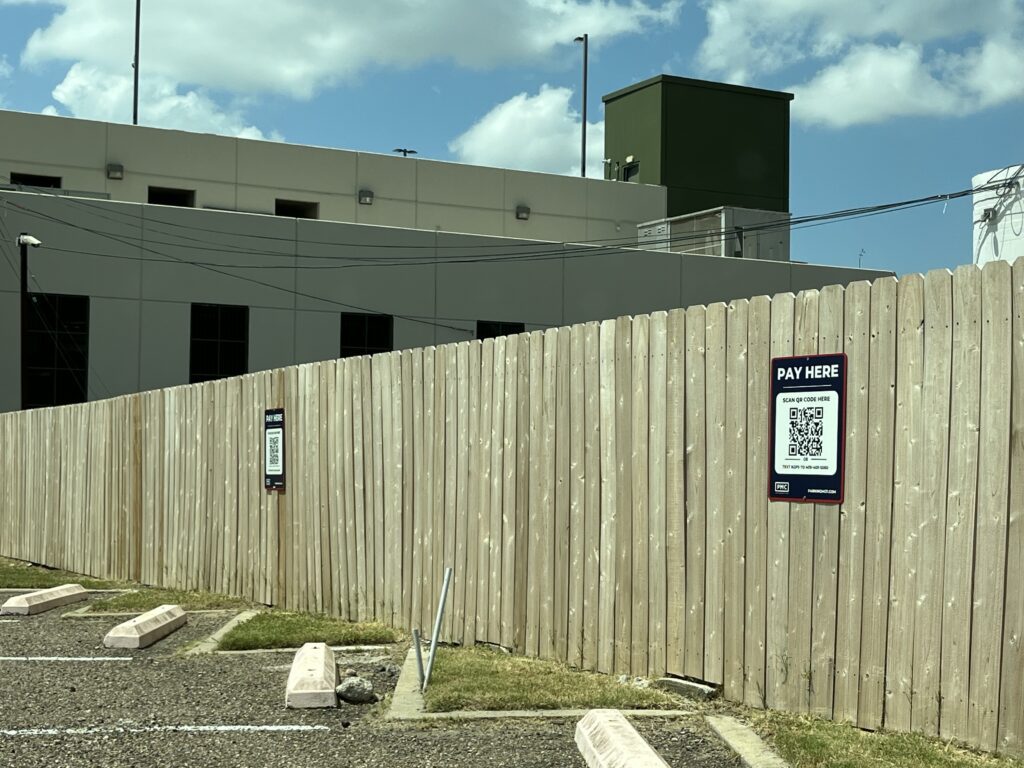 A wooden fence with two “Pay Here” QR code signs runs along a row of empty parking spaces in a lot next to a beige building under a partly cloudy sky.