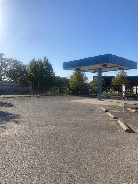 An empty parking lot with a blue-roofed canopy structure and parking bumpers under a clear, sunny sky. Trees and a fence line the background.