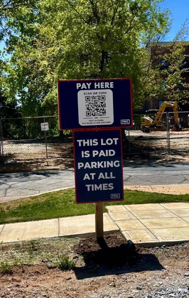 Two signs in front of a parking lot read: “PAY HERE” with a QR code, and “THIS LOT IS PAID PARKING AT ALL TIMES.” Trees, a sidewalk, and construction equipment are visible in the background.