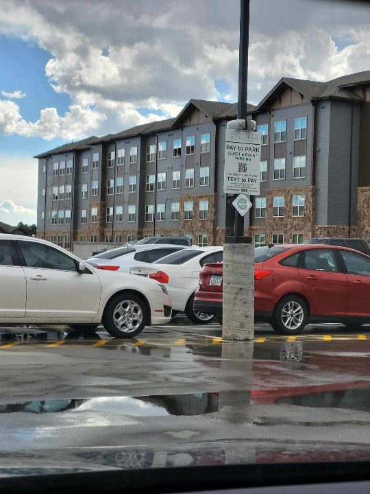 A parking lot with several parked cars in front of a multi-story apartment building. A sign on a pole instructs drivers to pay for parking. The pavement is wet, reflecting the cloudy sky above.