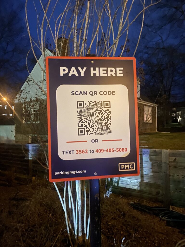 A sign at night reads "Pay Here" with a QR code and instructions to scan it or text "3562" to 409-405-5080 for payment. The sign is on a post near houses and trees.