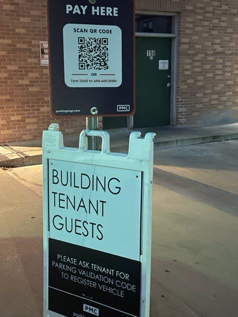 A sidewalk sign reads "Building Tenant Guests: Please ask tenant for parking validation code to register vehicle." Above it, a sign displays a QR code and instructions to pay for parking, attached to a post near a brick building.