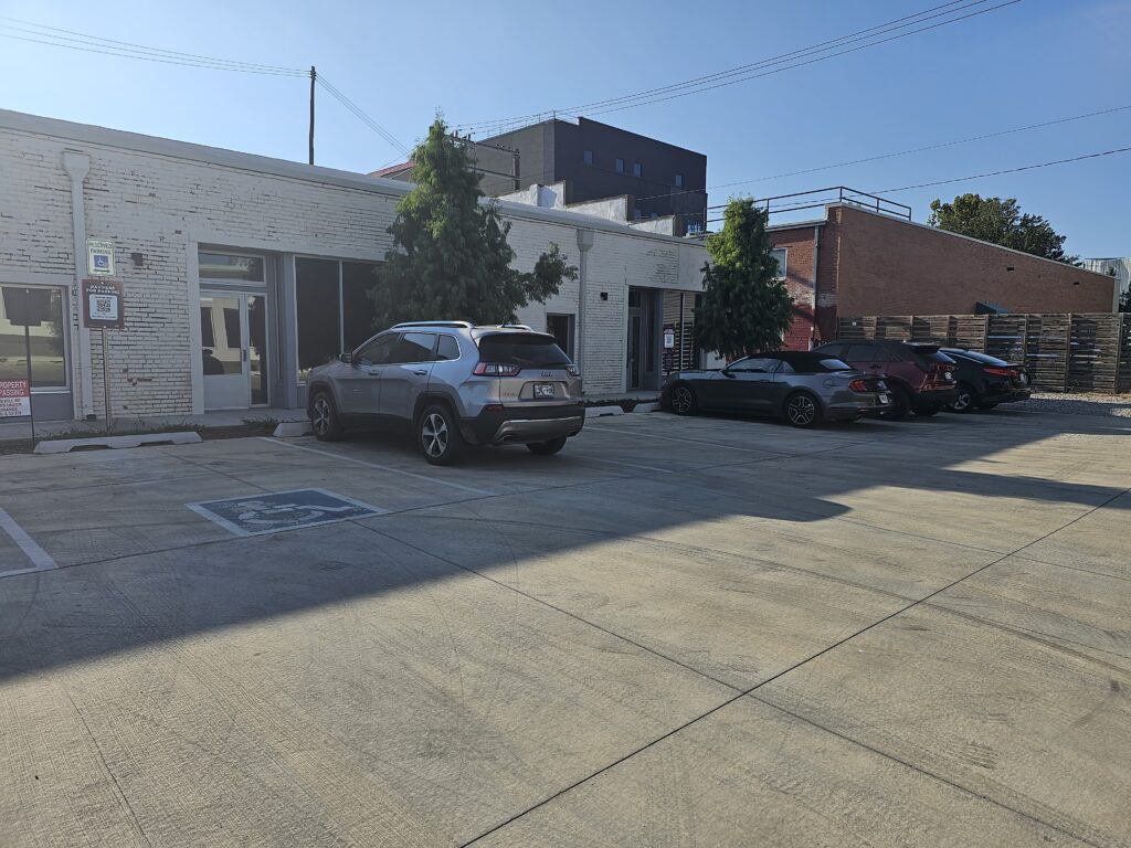 A small parking lot outside a light-colored brick building with four parked cars, including one in an accessible parking space. Trees line the building, and stacked pallets are visible in the background.