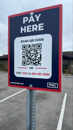 A parking lot sign reads "PAY HERE" with instructions to scan a QR code or text "YFZG" to 409-405-5080 for payment. The sign is mounted on a metal pole near empty parking spaces.