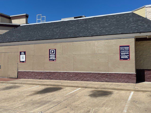 A beige building wall facing an empty parking lot with three signs about monthly parking rules and payment information; the sky is clear and blue above.