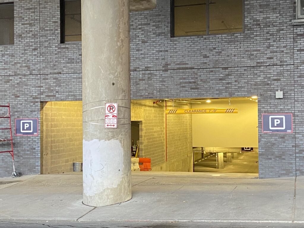 A parking garage entrance with a large concrete pillar and a No Parking sign in front. Two blue parking signs are on either side of the entrance, and the clearance bar inside reads "CLEARANCE 8'–0".