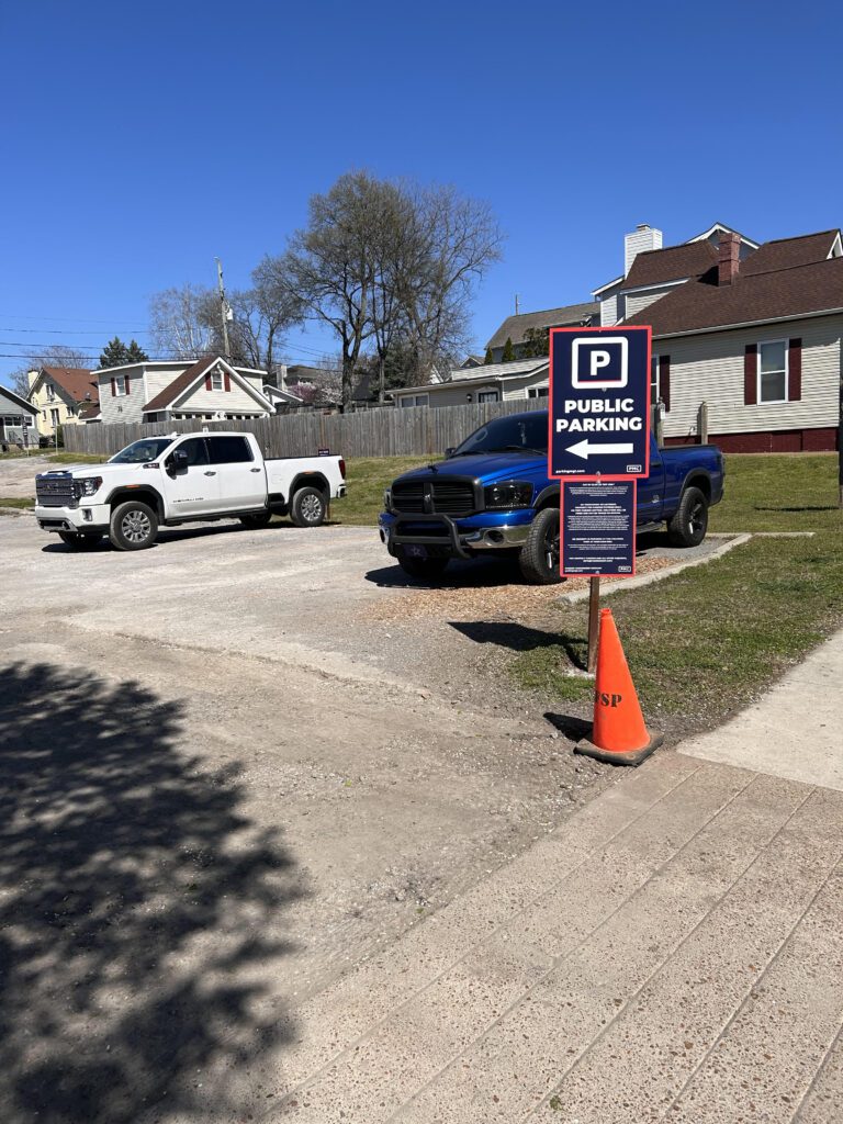 A small gravel parking lot with a white and a blue pickup truck parked. A sign reads "Public Parking." An orange traffic cone is placed near the sign. Houses and trees are visible in the background under a clear blue sky.
