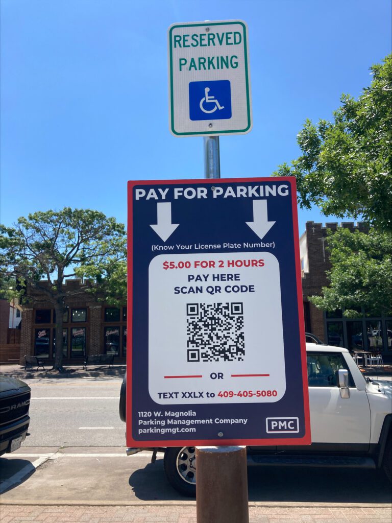 A sign designates a handicapped parking spot and displays a "Pay for Parking" instruction with a QR code, stating $5 for 2 hours. Parked cars and storefronts are visible in the background.