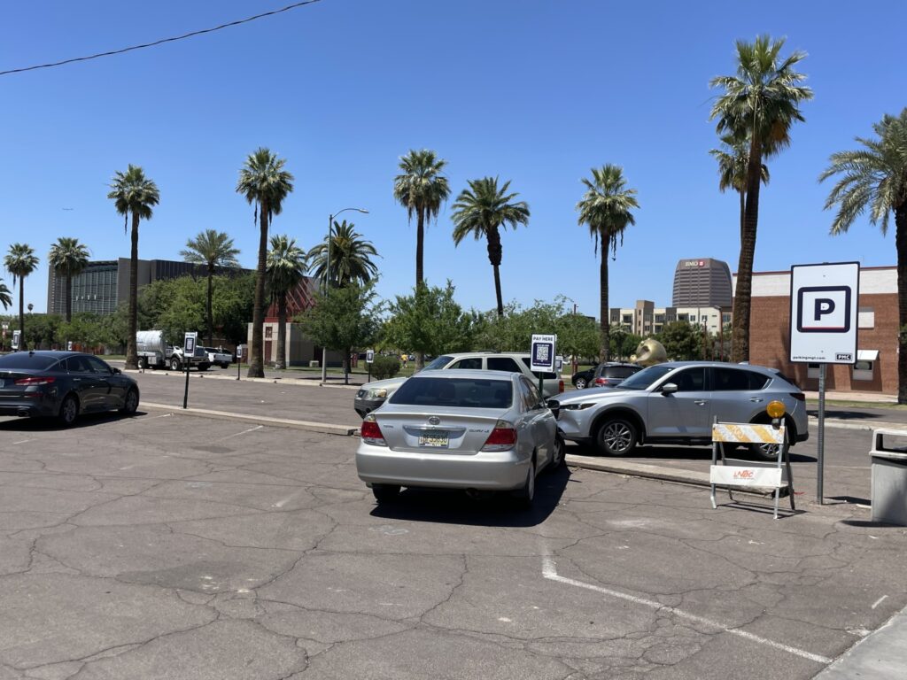 A PMC-owned parking lot with several parked cars, palm trees, and buildings in the background. A PMC parking sign is visible in the foreground.