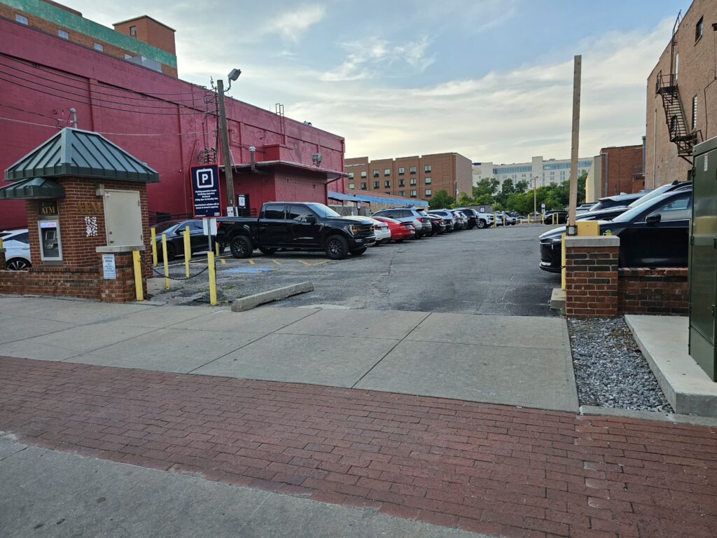 A parking lot filled with cars next to a sidewalk, with a small brick booth on the left and a large red building in the background. The sky is partly cloudy and several other buildings are visible.