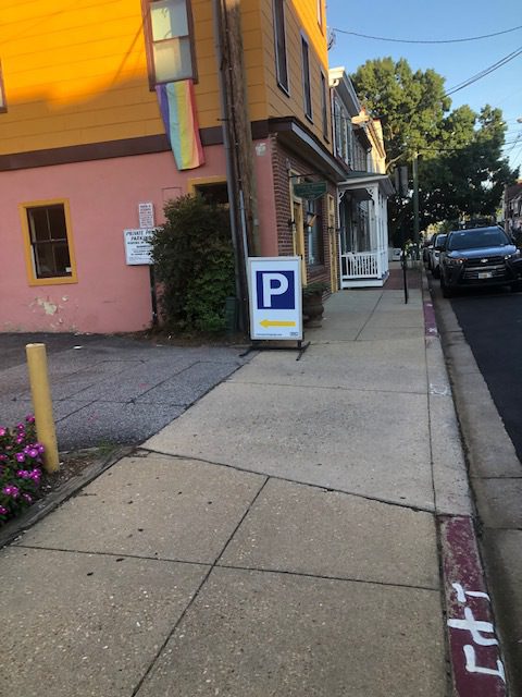 A sidewalk next to a yellow building with a rainbow flag. A sign with a "P" and a yellow arrow points toward parking. Cars are parked along the street lined with trees.