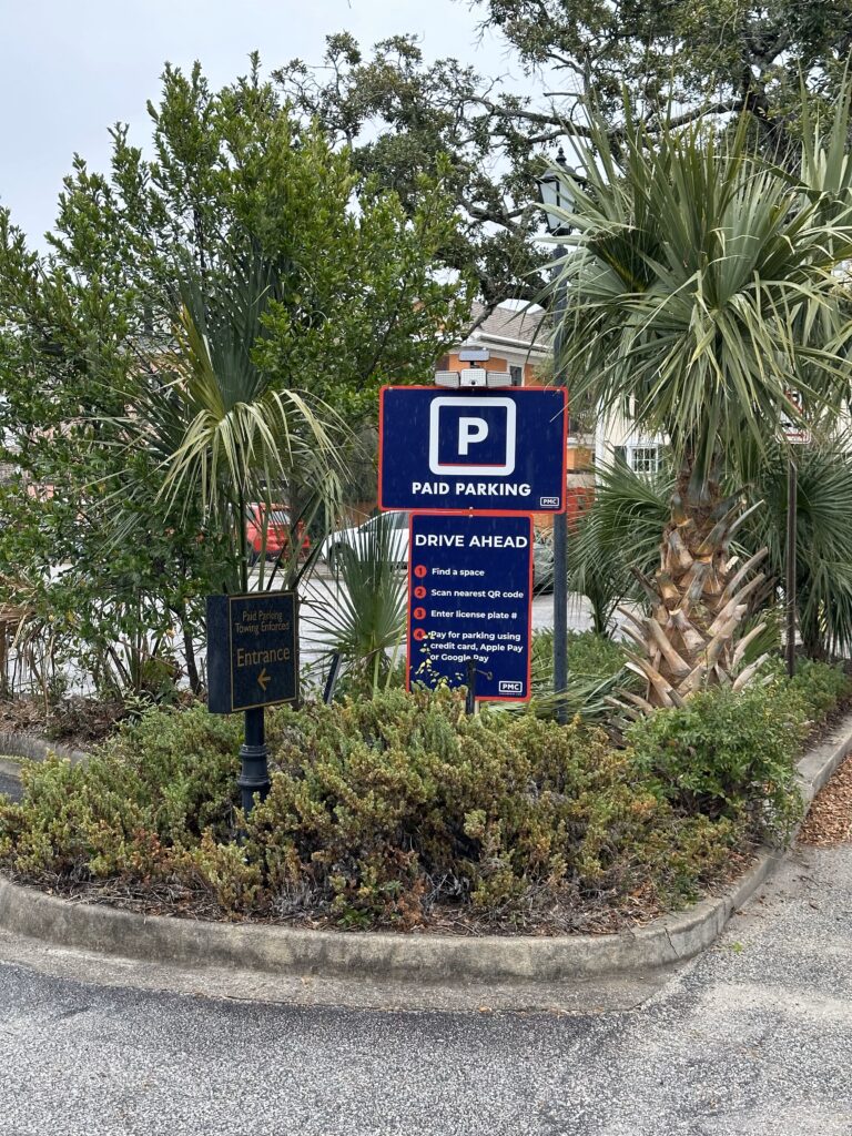 A sign surrounded by greenery displays "Paid Parking" and instructions for payment in a parking lot. Another smaller sign nearby indicates the entrance. Palm trees and plants are visible in the background.