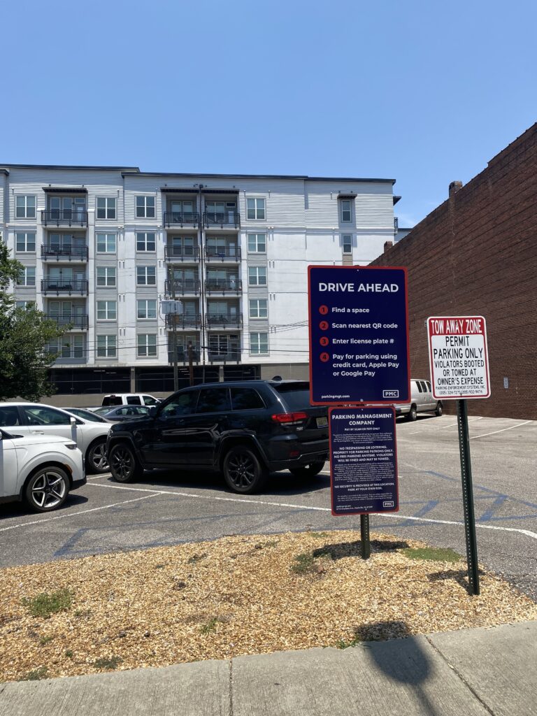 A parking lot with several cars parked, bordered by buildings. Signs display parking instructions, including QR code payment, and a notice for tow-away zone, permit parking only. A modern apartment building stands in the background.