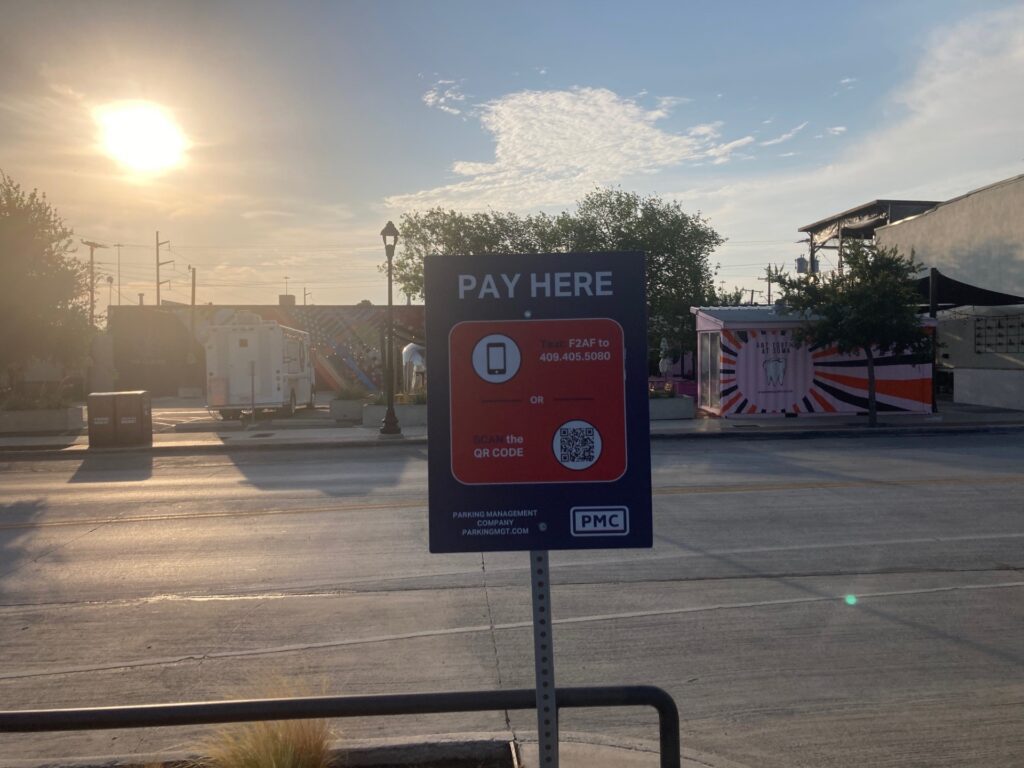 A red and blue “Pay Here” parking sign stands on a sidewalk in front of an empty street, with sunlight shining through trees and tents in the background under a partly cloudy sky.