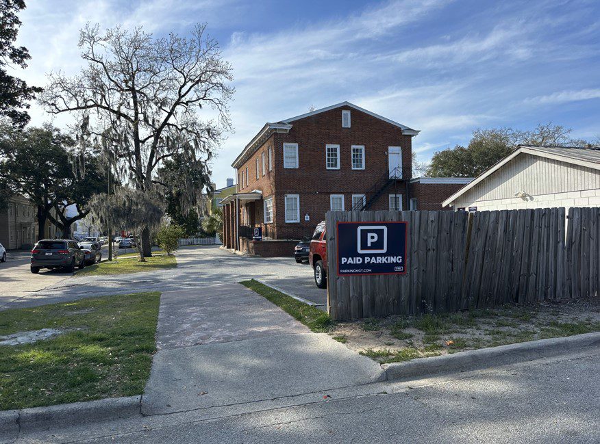 A small Parking Management Company parking lot next to a red brick building, with a sign on a wooden fence that reads "Paid Parking" with the PMC logo above it. Several cars are parked under a clear blue sky with scattered clouds.