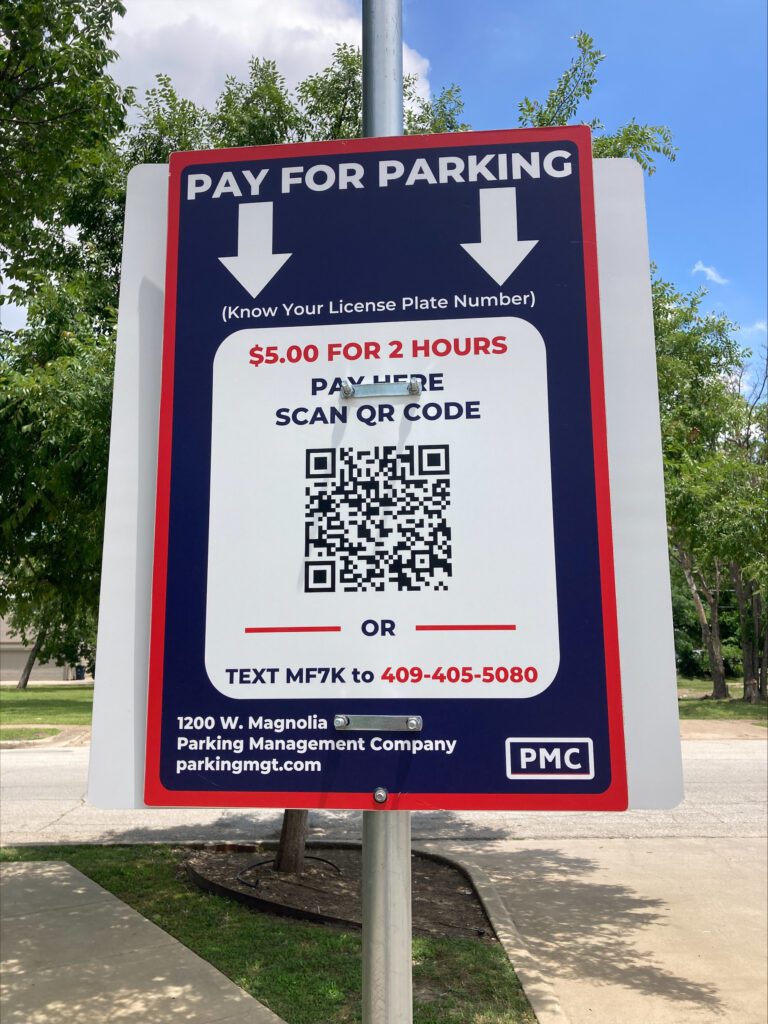 A sign on a metal pole reads "Pay for Parking" with instructions to scan a QR code or text MF7K to a phone number. Parking costs $5 for 2 hours. Trees, grass, and a sidewalk are in the background.