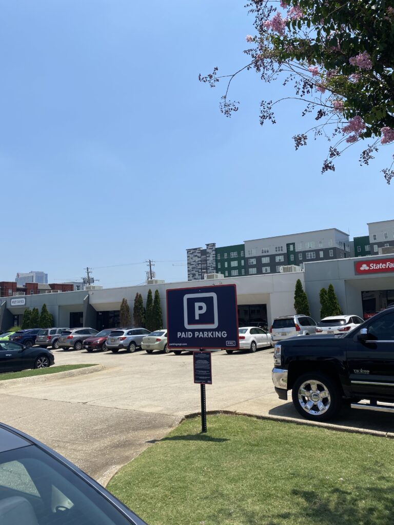 A parking lot with several parked cars, a sign in the foreground that says "PAID PARKING," and shops and apartment buildings in the background under a clear blue sky.