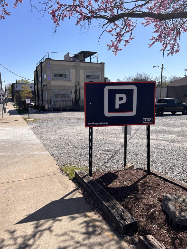 A large "P" sign marks a gravel parking lot next to a sidewalk on a sunny day, with a multi-story brick building and blooming tree branches in the background.