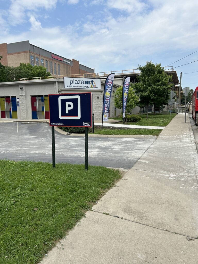 A parking lot with a large "P" parking sign in front of a building with colorful windows. Two flags reading "Now Open" stand by the entrance, and a hospital building is visible in the background.