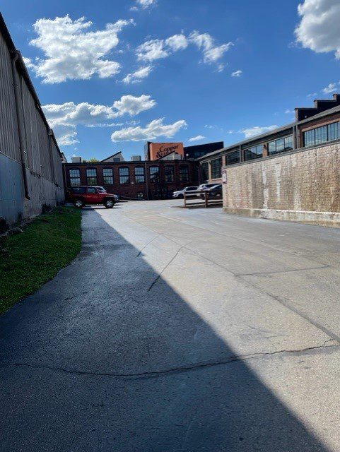 A paved alleyway runs between two industrial buildings under a blue sky with scattered clouds. A red car and a small white structure are visible in the distance near a building labeled “Sonder.”.