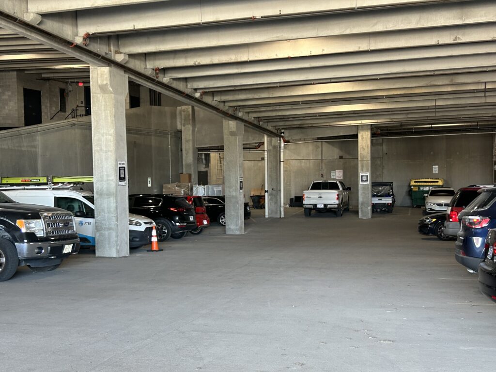 Indoor parking garage with concrete pillars and beams, several parked cars, trucks, and a van. Some orange cones and trash bins are visible in the background, along with signs on the pillars. The lighting is natural.