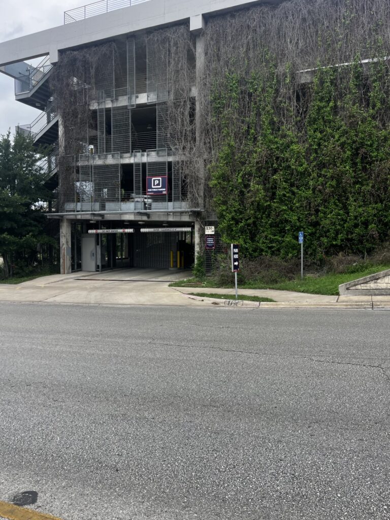 A multilevel parking garage covered in vines on the right side, with an entrance visible on the ground floor and a parking sign nearby. The street is empty in the foreground.