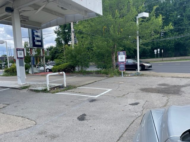 An empty, cracked parking lot at a gas station with faded markings; gas price signs are visible but blank. Trees and a black car driving on a street are in the background.
