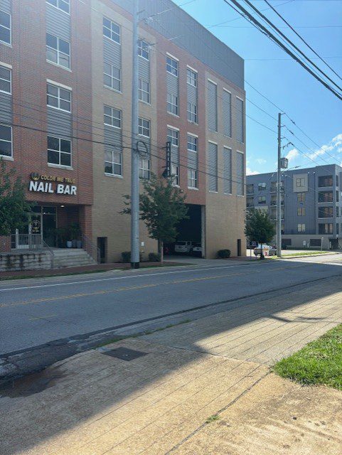A street view showing a brick building with a "Color Me TNC Nail Bar" sign on the left. There is an open garage entrance with a car inside, trees, power lines, and other modern buildings in the background.