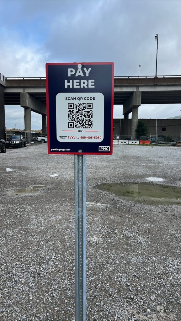 A sign in a gravel parking lot reads "Pay Here." It includes a QR code for payment, a phone number for text payment, and instructions. An overpass bridge is visible in the background under a cloudy sky.