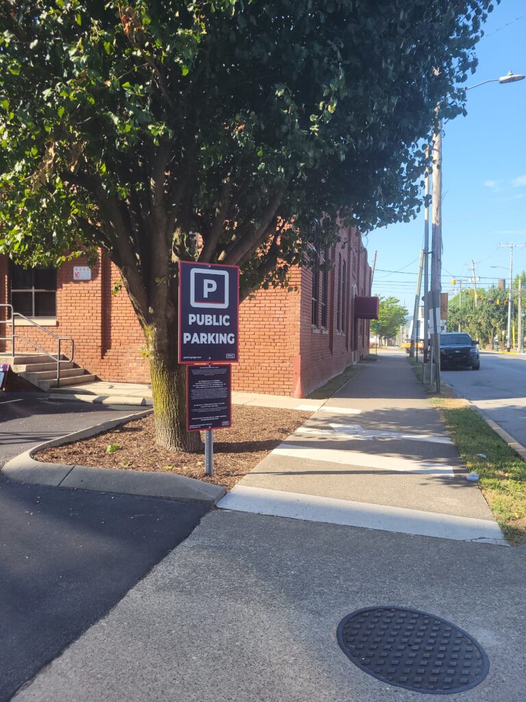 A "Public Parking" sign stands in front of a large tree by the sidewalk next to a red brick building on a sunny day. The street and some utility poles are visible in the background.