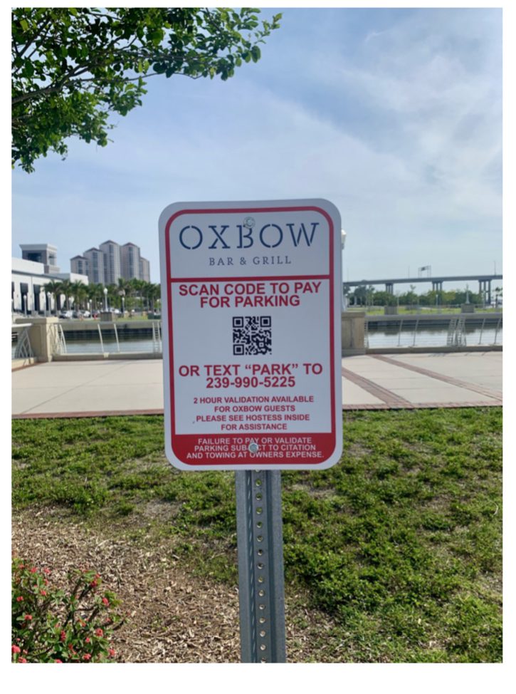 A sign for Oxbow Bar & Grill instructs visitors to scan a QR code or text "PARK" to a phone number to pay for parking. The background includes a walkway, waterfront, and buildings.