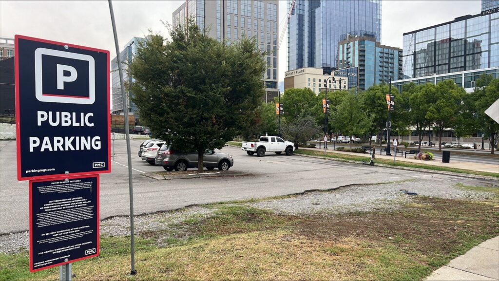 A public parking lot in a city with several parked cars, a large "Public Parking" sign in the foreground, and tall buildings with glass windows in the background. Trees line the street near the lot.