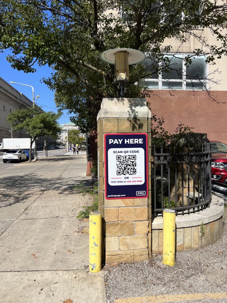 A "Pay Here" parking sign with a QR code is posted on a stone pillar next to a sidewalk and street on a sunny day. Trees, yellow posts, and a building are visible in the background.