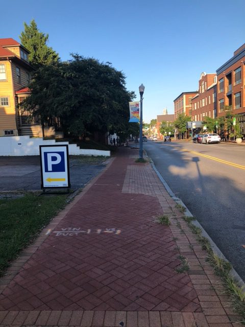 A brick sidewalk runs alongside a street lined with buildings. A sign with a "P" and a yellow arrow points left toward parking. Trees provide some shade, and cars are parked along the street under a clear blue sky.