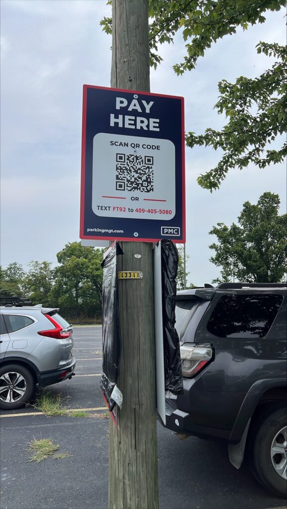 A parking payment sign on a wooden pole instructs users to scan a QR code or text "FT92" to a listed number. Trees and parked cars are visible in the background.