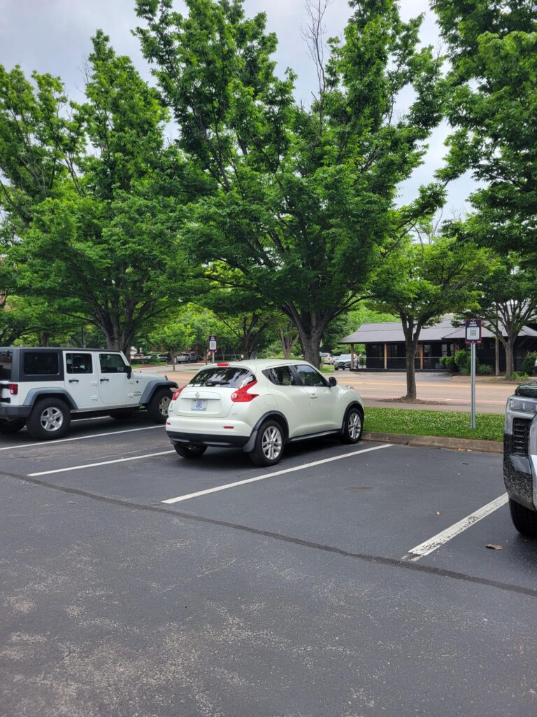A white compact SUV is parked in a parking lot next to a white Jeep. The lot is bordered by lush green trees, and a building is visible in the background under a cloudy sky.