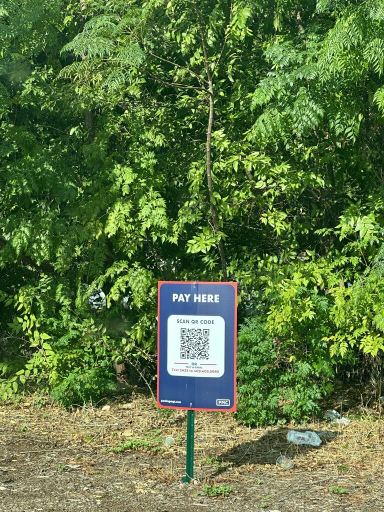 A "Pay Here" sign with a QR code stands in front of dense green trees and bushes. The ground is covered with dry leaves and debris. The sign provides payment instructions for a parking or service area.