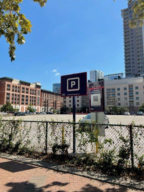 A parking lot with a few cars, surrounded by tall buildings. A parking sign and payment QR code are visible in the foreground, with a chain-link fence and leafy tree branches overhead.