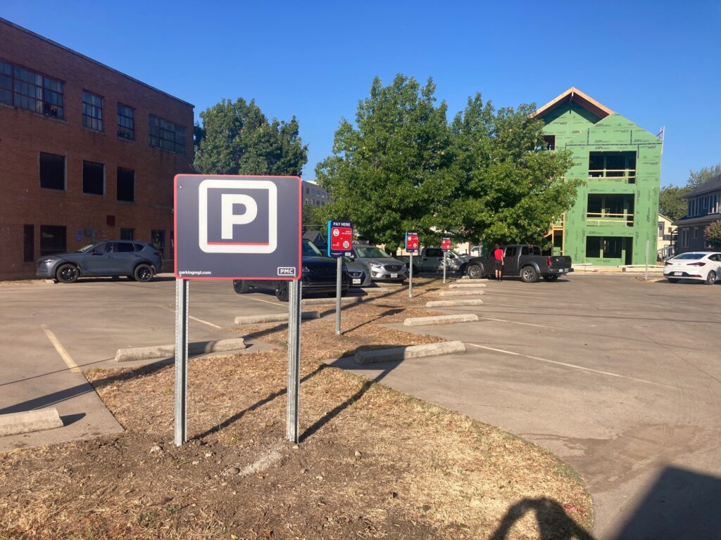 An outdoor parking lot with several parked cars, visible "P" parking signs, a brick building on the left, and a partially constructed green building in the background on a sunny day.