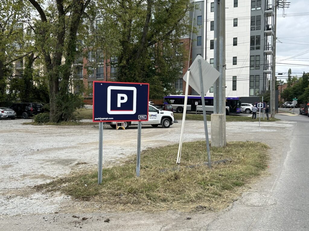 A gravel parking lot with a prominent parking sign in the foreground, bordered by trees and grass. Several parked cars and a white building with multiple floors are visible in the background.