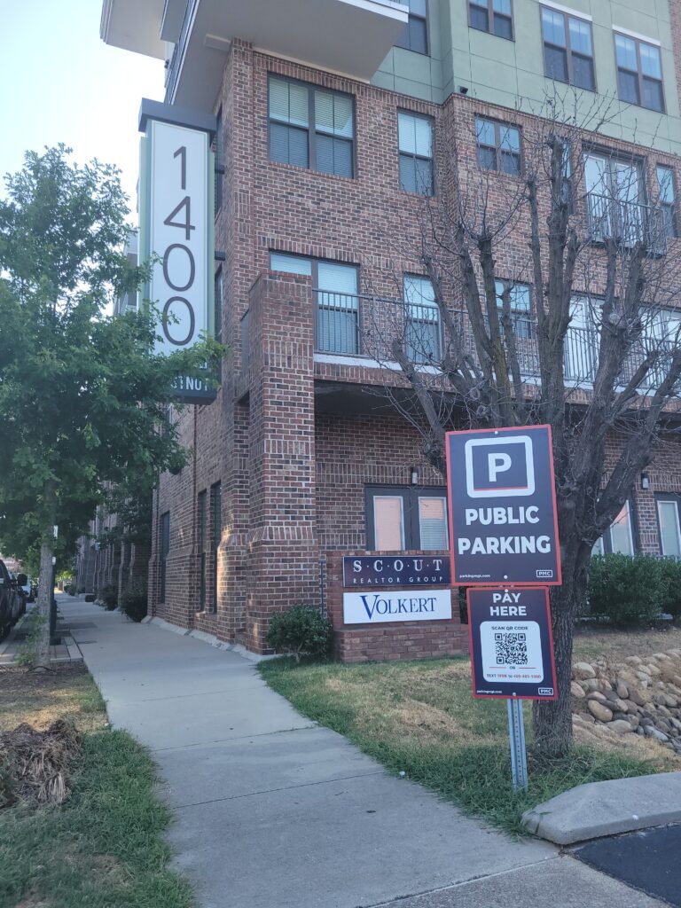 A brick apartment building with the address "1400" displayed vertically, a sidewalk in front, and a sign for public parking with a QR code for payment. A tree and some shrubbery are visible beside the building.