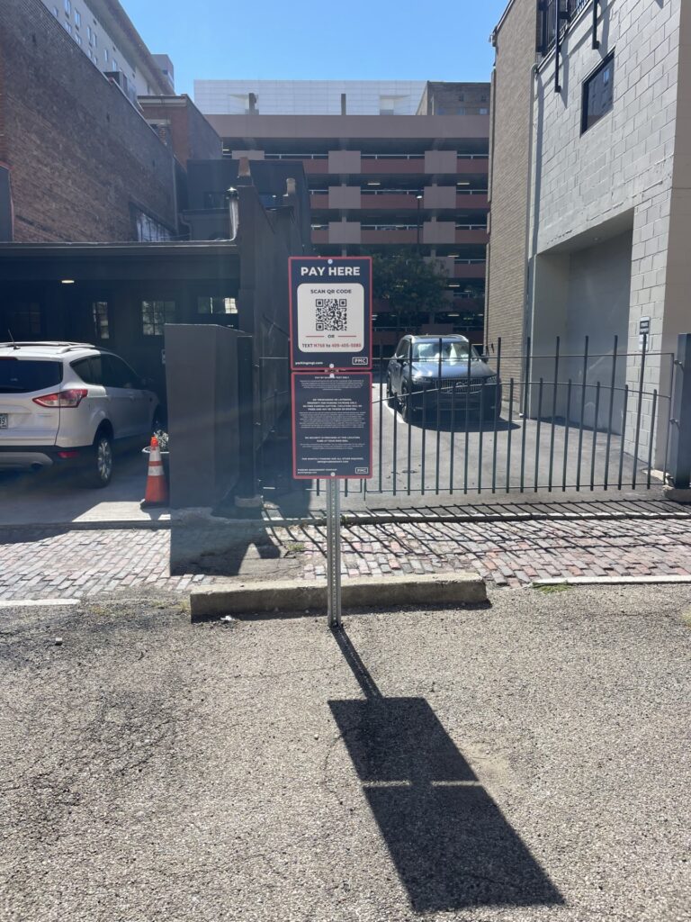 A metal sign in a parking lot reads "Pay Here" with a QR code and payment instructions. Cars are parked nearby, and buildings surround the area. The sun casts a shadow of the sign on the ground.