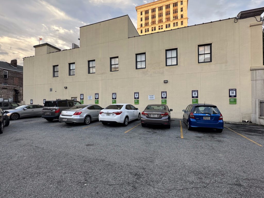 Parking Management Company-owned parking lot with six parked cars in front of a beige building. PMC signs are posted on the wall with QR codes for digital payment, and a tall, ornate building is visible in the background under a partly cloudy sky.