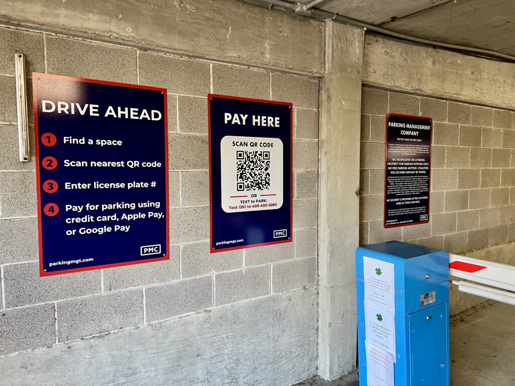 Three PMC signs are mounted on a concrete wall in a parking garage. The sign on the left has instructions for paying for parking, the middle sign has a QR code for digital payment, and the sign on the right has parking regulations and ownership details. A blue payment machine and parking gate are in the foreground.