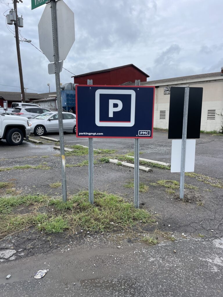 A blue and white “P” parking sign stands on a patch of grass in a lot with several cars parked nearby. Buildings and a cloudy sky are in the background.