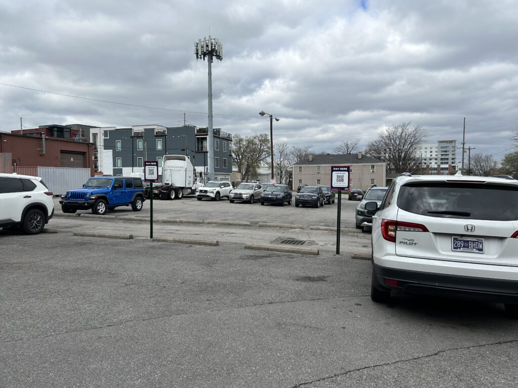A mostly empty urban parking lot with several parked cars, a few small buildings in the background, and a large cell tower. The sky is overcast with gray clouds. Signs are visible near the parked vehicles.