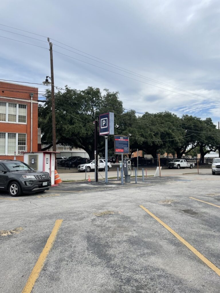A mostly empty outdoor parking lot with yellow lines, payment machines, and a parking sign. A few parked cars, a brick building, and large green trees are in the background under a cloudy sky.