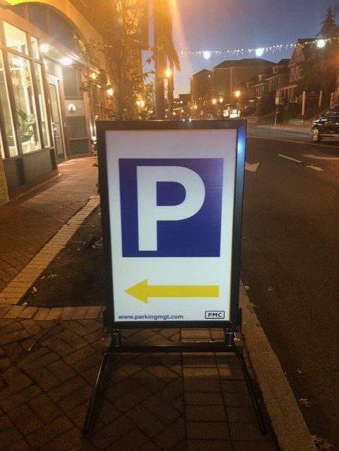 A sidewalk sign with a large blue "P" for parking and a yellow arrow pointing left, directing drivers toward parking. The sign is on a brick sidewalk along a street at dusk with lights overhead.